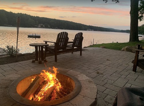 A cozy lakeside scene at sunset, featuring a fire pit, wooden chairs, and calm water in the background.