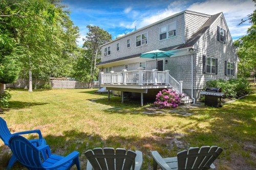 A backyard view of a house with a deck, green lawn, and colorful flowers, featuring blue chairs and a grill.