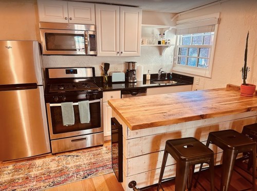 Modern kitchen with stainless steel appliances, a wooden island, and bar stools, featuring a cozy rug and natural light.