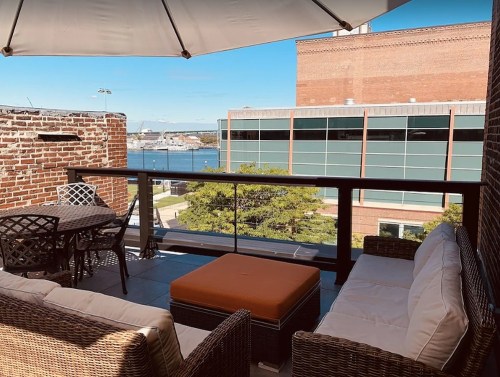 A cozy balcony with wicker furniture, an orange ottoman, and a view of a waterfront and buildings under a clear blue sky.