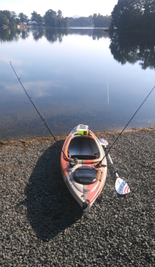 A kayak with fishing rods rests on a gravel shore by a calm lake, surrounded by trees and clear blue skies.