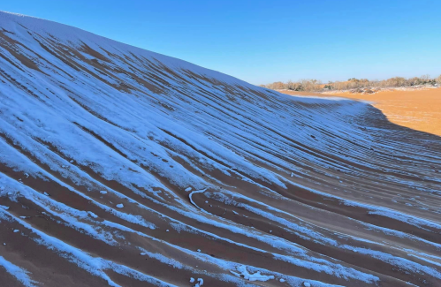 A sandy dune with snow patches, under a clear blue sky, showcasing contrasting textures and colors.