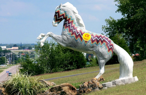A colorful, patterned horse statue rearing up on a grassy hill near a road, with trees and a blue sky in the background.