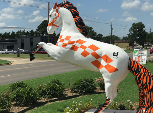 A rearing horse statue painted white with orange checkers and black stripes, set against a blue sky and green grass.