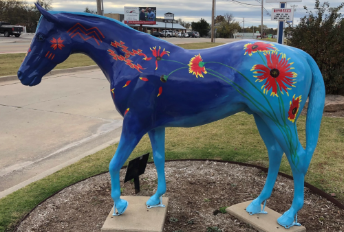 A blue horse statue decorated with colorful flowers, standing on a grassy area near a road.