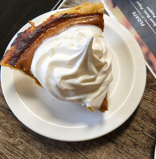 A slice of pie topped with a swirl of whipped cream on a white plate, resting on a wooden table.