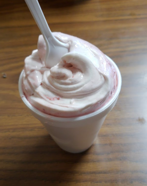 A cup of soft-serve ice cream swirled with pink strawberry sauce and topped with a plastic spoon.