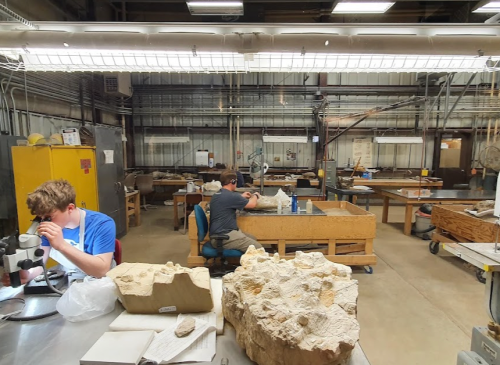 Two students work in a spacious lab filled with tools and materials, one using a microscope and the other focused on a project.