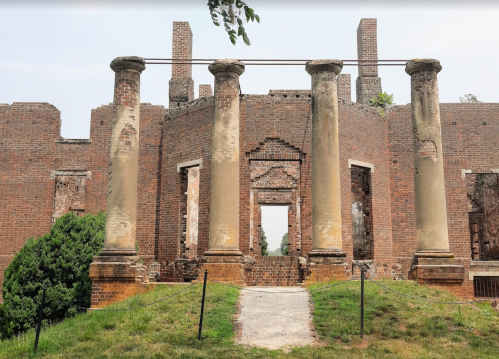 Ruins of a brick building with tall columns, surrounded by greenery and a gravel path leading to the entrance.