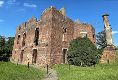 Ruins of a brick building with broken windows, surrounded by greenery and a stone chimney, under a clear blue sky.