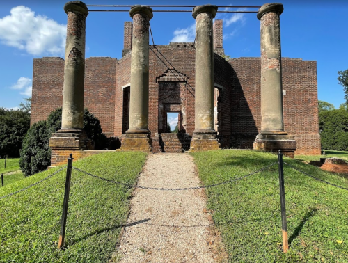 Ruins of a brick building with four tall columns, surrounded by grass and trees, under a blue sky with clouds.