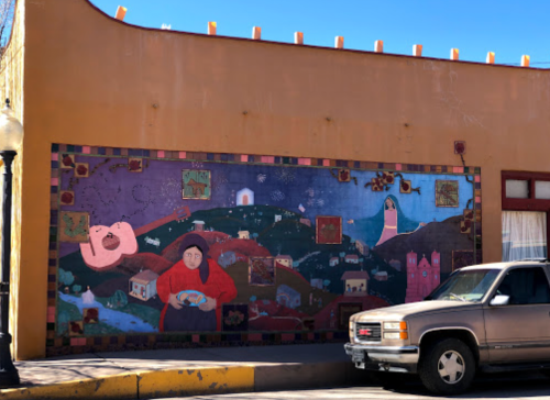 A colorful mural depicting a woman with a bowl, a guitar, and various houses on a hillside, beside a parked truck.