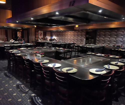 A spacious restaurant interior with multiple tables set for dining, featuring a central cooking area and dark wood decor.