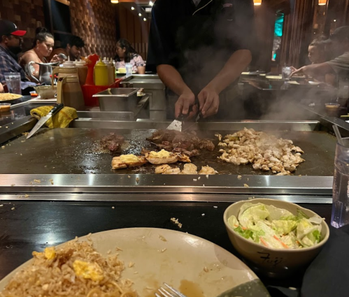 A chef cooks meat and vegetables on a grill, with a plate of fried rice and a salad in the foreground.