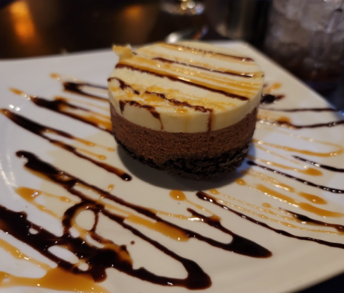 A slice of layered chocolate and vanilla cheesecake drizzled with chocolate and caramel sauces on a white plate.