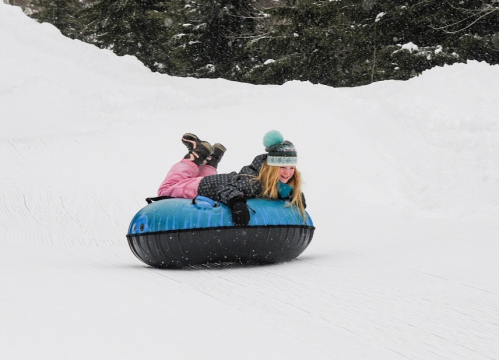 A girl in winter attire joyfully rides a blue and black snow tube down a snowy slope. Snow is falling around her.