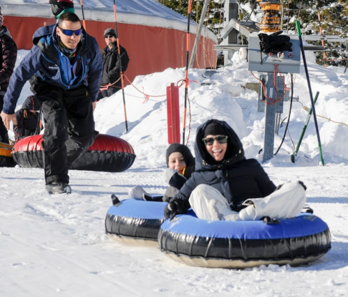 Two people in snow tubes smile as they slide down a snowy hill, while an instructor assists from behind.