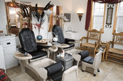 A cozy nail salon featuring two black pedicure chairs, a small table with nail supplies, and wooden chairs in a bright room.