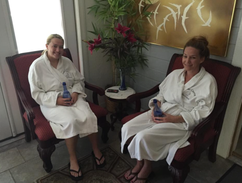Two women in white robes sit in armchairs, holding water bottles, surrounded by plants and decorative art.