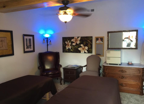 A cozy room with two massage tables, a chair, and decorative artwork on the walls, featuring soft lighting.