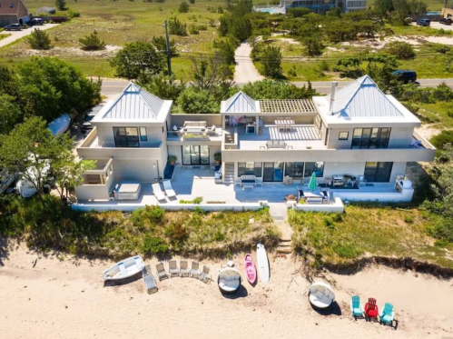 Aerial view of a modern beach house with a sandy shore, surrounded by greenery and colorful beach chairs.