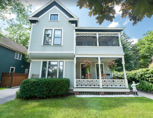 A charming two-story house with a porch, surrounded by greenery and flowers, on a sunny day.