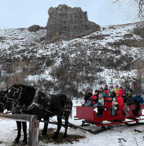 A group of people in winter attire sits on a red sleigh pulled by a horse, with snowy hills in the background.