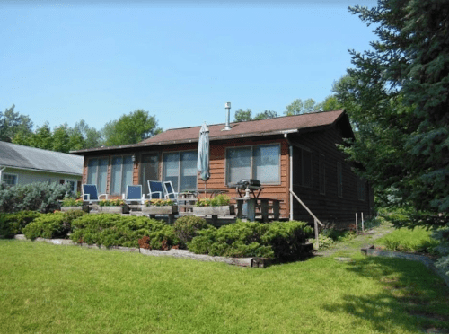 A rustic wooden house with a deck, surrounded by greenery and trees on a sunny day.