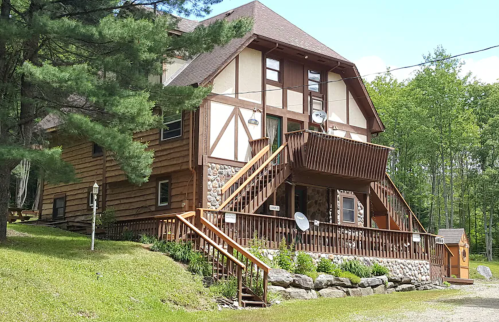 A large, rustic wooden house with a stone foundation, featuring a front porch and surrounded by trees.
