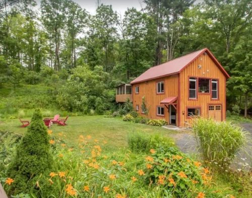 A charming wooden house with a red roof, surrounded by greenery and orange flowers, set in a peaceful outdoor area.