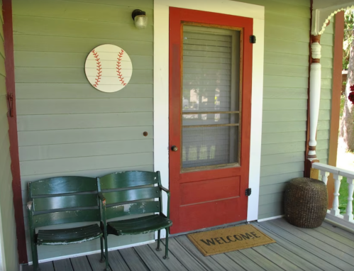 A welcoming porch with a red door, green benches, a baseball decoration, and a "WELCOME" mat.