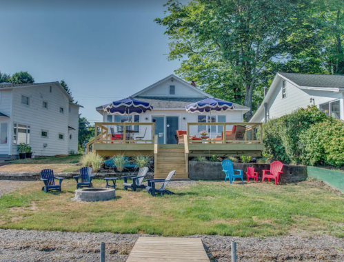 A cozy house with a deck, surrounded by colorful chairs and greenery, on a sunny day.