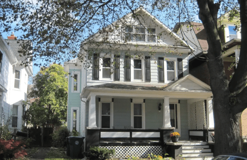 A two-story house with a light blue exterior, white trim, and a porch, surrounded by trees and greenery.