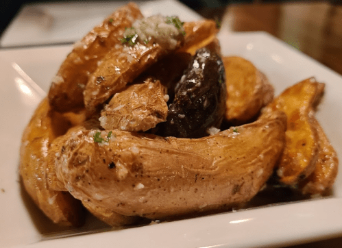 A close-up of golden-brown roasted potatoes garnished with herbs and a sprinkle of seasoning in a white dish.
