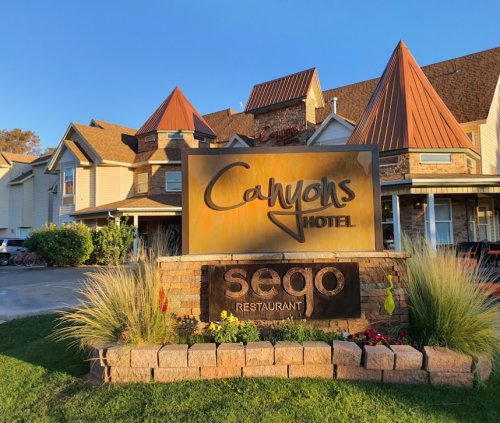 Sign for Canyons Hotel and Sego Restaurant, featuring a stone base and surrounded by greenery and colorful flowers.