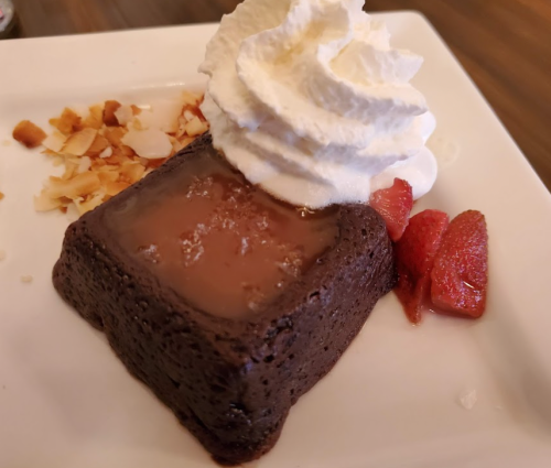 A square chocolate dessert topped with whipped cream, served with strawberries and coconut flakes on a white plate.