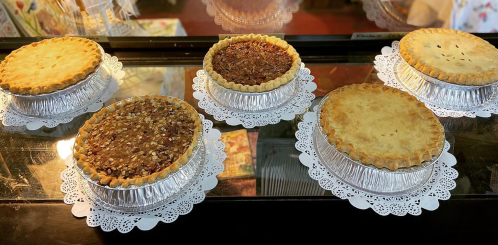 Five pies in silver foil pans on lace doilies, featuring a variety of fillings, displayed in a glass case.