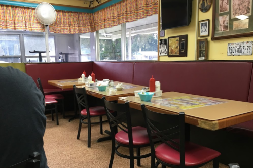 A cozy diner interior with red booths, tables, and condiments on the tables, featuring colorful decor and a fan.