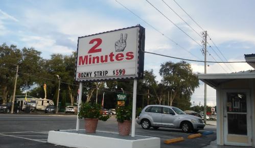 Sign for "2 Minutes" featuring a peace hand symbol, advertising an 8 oz. NY strip for $5.99, with a parking lot in view.