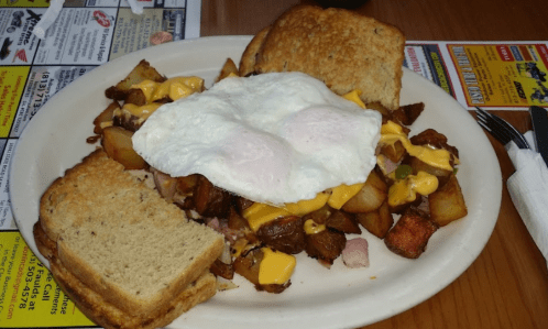 A plate of breakfast featuring crispy potatoes, cheese, ham, two fried eggs, and toasted bread on the side.