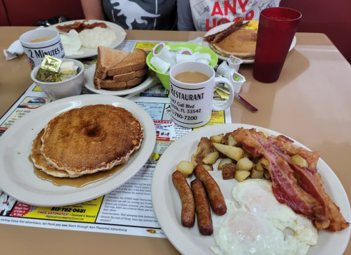 A breakfast spread featuring pancakes, eggs, bacon, sausage, potatoes, toast, and coffee on a diner table.