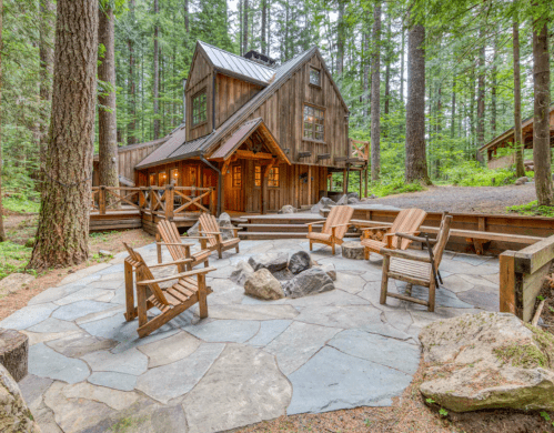 A cozy cabin surrounded by trees, featuring a stone patio with wooden chairs and a fire pit.