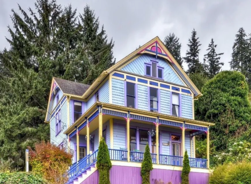 A colorful Victorian-style house with purple and blue siding, surrounded by greenery and tall trees.