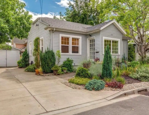 A charming gray house with a landscaped garden and driveway, surrounded by trees and shrubs.