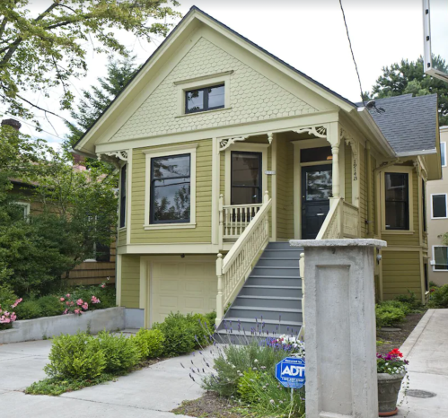 A charming yellow house with a front porch, stairs, and landscaped garden, set in a residential area.