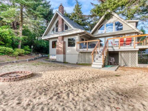 A modern house with large windows, surrounded by trees, and a sandy area with a fire pit in the foreground.