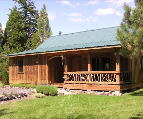 A rustic wooden cabin with a green metal roof, surrounded by trees and a landscaped yard.