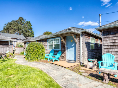 A cozy cottage with a blue door, surrounded by green grass and blue chairs, under a clear blue sky.