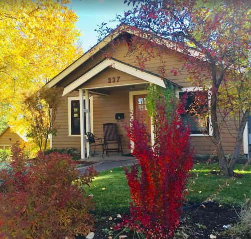 A cozy brown house with a porch, surrounded by vibrant autumn foliage and a striking red bush in the foreground.