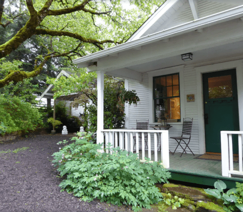 A charming white house with a porch, surrounded by greenery and a gravel path, featuring a small table and chairs.